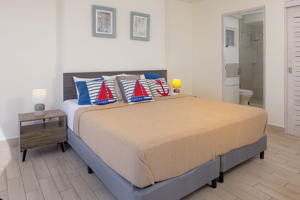 Een keurig opgemaakt bed met een beige deken, een nachtkastje met een lamp en een lijst met een foto aan de muur.