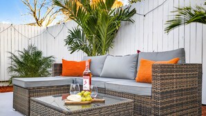 A wicker sofa with orange cushions, a glass coffee table with a bottle of wine and grapes, and a white fence in the background. - Family Luxury Escape Heated Pool, Spa, Mini Golf, Arcade (Largo)