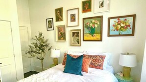 Habitación de hotel con una cama grande, varios almohadones, una mesita de noche con lámpara, una pequeña mesa con lámpara y varias obras de arte enmarcadas en la pared.