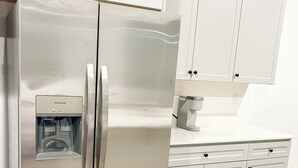 Un réfrigérateur en acier inoxydable doté d’un distributeur d’eau, de meubles blancs et d’un plan de travail sur lequel se trouve une machine à café.