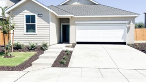 Une maison de plain-pied avec un garage, une petite cour avant agrémentée d’un arbre et de quelques arbustes, et un ciel dégagé.