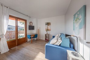 A living room with a blue sofa, a TV, a desk with a chair, and a sliding glass door leading to an outdoor area. - Le Fabre Azur - 600 m de la plage, parking (Saint-Aygulf)