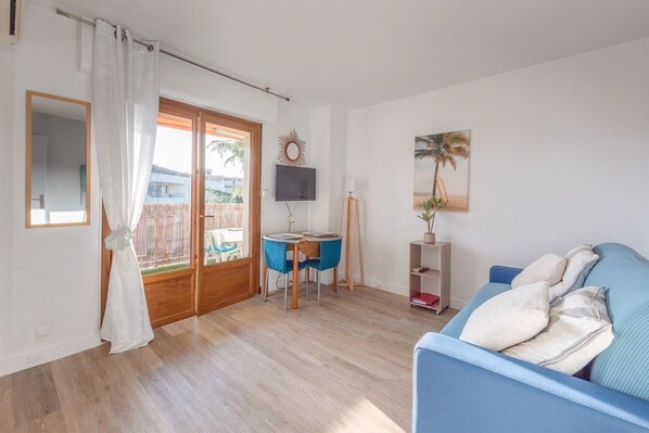 A living room with a blue sofa, a small table with chairs, a TV mounted on the wall, and a large window with curtains. - Le Fabre Azur - 600 m de la plage, parking (Saint-Aygulf)