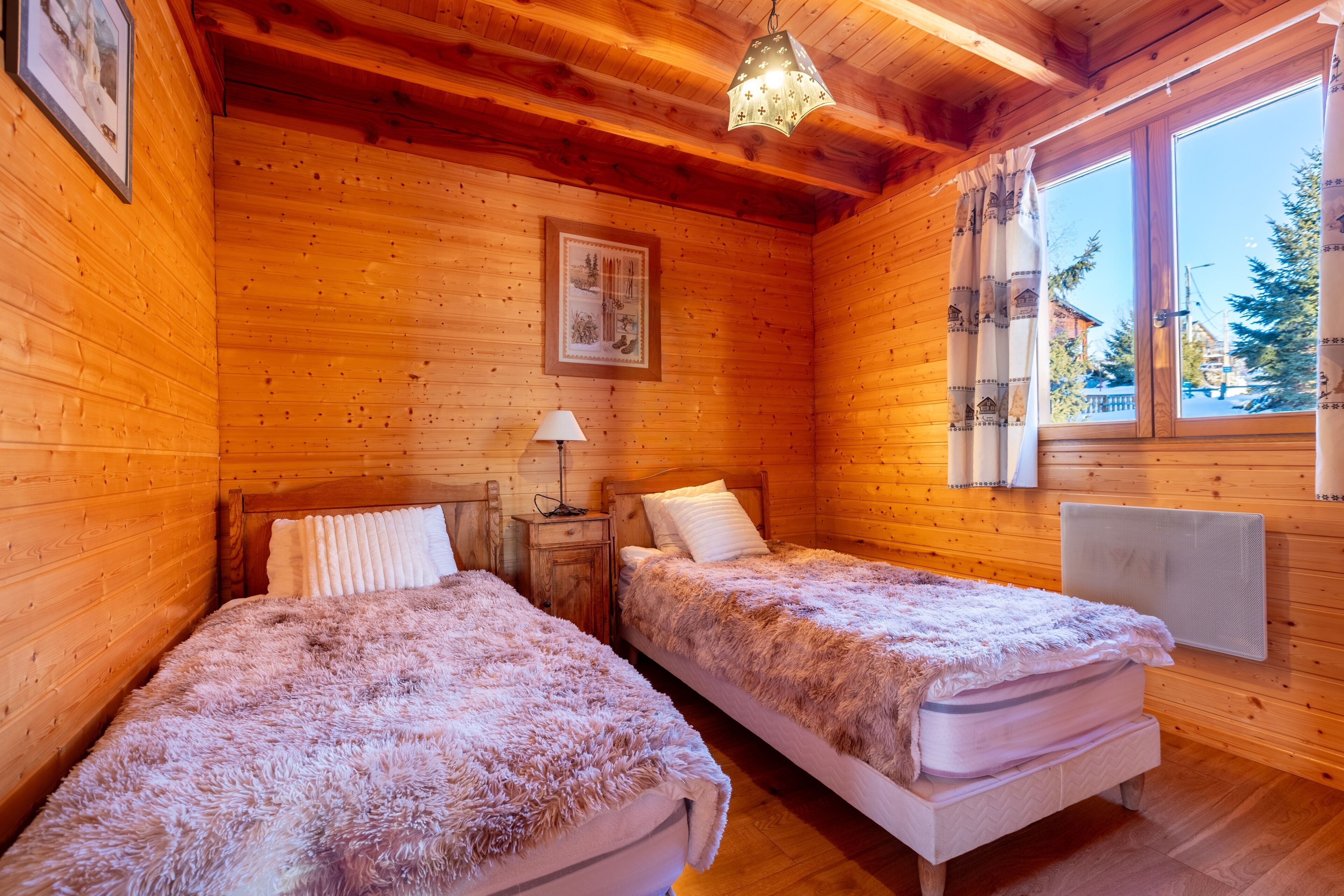 A wooden cabin room with two beds, a nightstand, a lamp, and a window with curtains.
