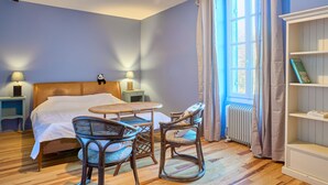 A bedroom with a bed, two wicker chairs, a small table, a nightstand with a lamp, a bookshelf, and a window with curtains. - Château Tour Chapoux - Piscine privée (Saint-Antoine-du-Queyret)