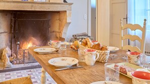 A dining table set with plates, glasses, and a basket of bread. A fireplace with a visible fire is in the background. - Château Tour Chapoux - Piscine privée (Saint-Antoine-du-Queyret)