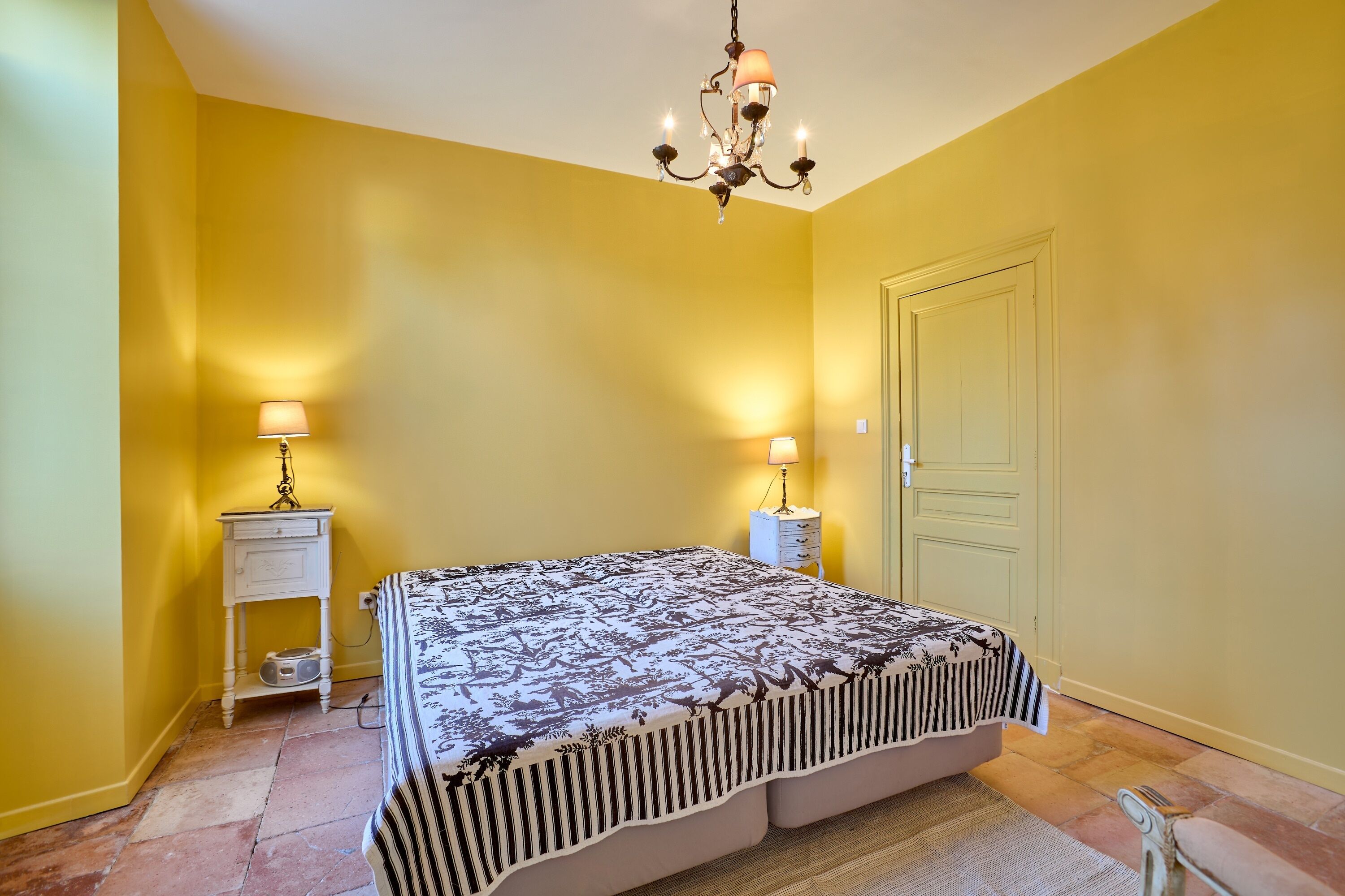 A bedroom with a bed, two bedside tables, a chandelier, and a door.