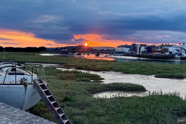 Orilla de río con barcos, un puente y edificios al fondo durante el atardecer.