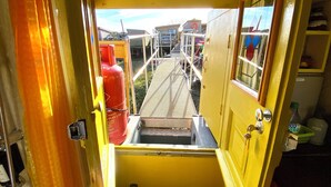 A narrow corridor with a metal grate floor, leading to an outdoor area with a red gas cylinder and a metal staircase.