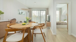 Una sala de estar moderna con una mesa de comedor redonda de madera, sillas blancas acolchadas, un ventanal con cortinas, un televisor de pantalla plana y una cama con cubrecama color beige al fondo.