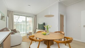 Una sala de estar moderna con una mesa de comedor redonda de madera, sillas blancas acolchadas, un televisor de pantalla plana y un amplio ventanal con cortinas.