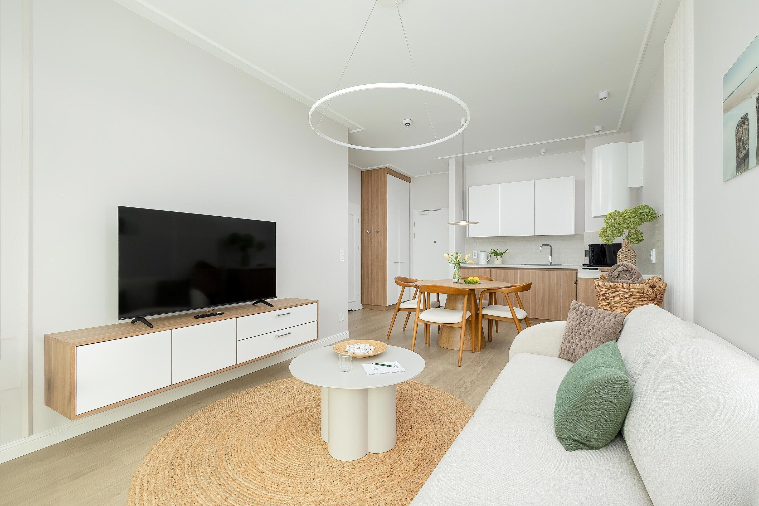 A modern living room with a flat-screen TV, a white sofa, a round coffee table, and a dining area with wooden furniture.