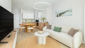 Una sala moderna con un sofá, una mesa de centro y un televisor. Se observa un comedor con una mesa y sillas. La cocina es visible al fondo.