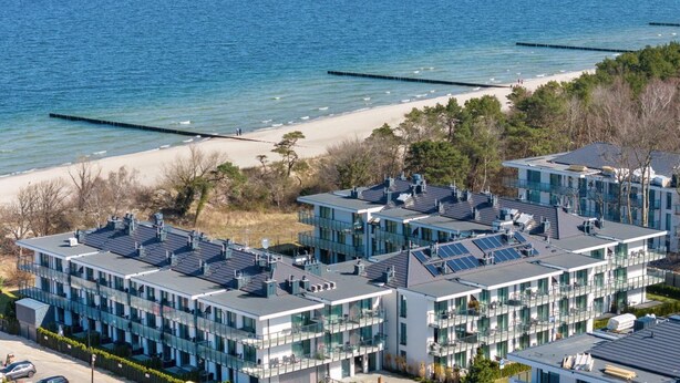 Un edificio moderno de varios pisos con amplios ventanales y paneles solares en el techo, situado cerca de una playa con arena y un muelle.