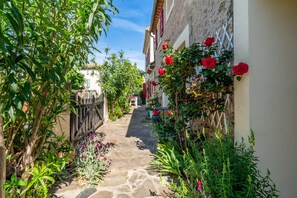 A narrow stone pathway flanked by lush greenery and red flowers, leading to a white building with a red door. - “Galet” Room with Shared Terrace, Wi-Fi, and Air Conditioning (Colombiers)