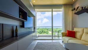Ein modernes Wohnzimmer mit einer Couch, einem Couchtisch und einem an der Wand montierten Fernseher. Es gibt einen Balkon mit Blick auf die Grünflächen.