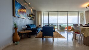Ein modernes Wohnzimmer mit großem Fenster, einem blauen Sofa, einem Essbereich mit Tisch und Stühlen sowie einem Gemälde an der Wand.