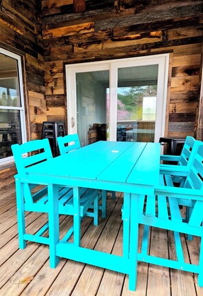 Une table et des chaises en bois turquoise sont disposées sur une terrasse en bois, et l’intérieur de la cabane a un style rustique.