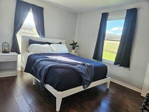 Une chambre à coucher avec un lit, des tables de chevet blanches, une plante et une vue sur l’extérieur.