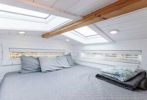 A bedroom with a skylight, a bed with grey bedding, and a view of the outside. - Pet-Friendly Zion Tiny Home • Private Hot Tub (Apple Valley)