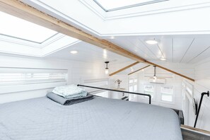 Une chambre avec une lucarne, un lit recouvert d’une courtepointe, un luminaire suspendu et une vue sur l’extérieur.