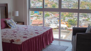 A bedroom with a large window, a bed with a floral bedspread, a chair, and a nightstand with a lamp. - Apartment with a beautiful view of the Bay (Acapulco de Juárez)