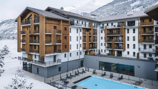 Um hotel moderno de vários andares com área de piscina, rodeado por montanhas nevadas.
