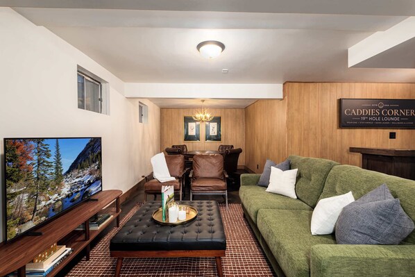 A modern living room with a large flat-screen TV, a green sofa, a black ottoman, and a wooden dining table with chairs. - Arvada Retreat | Garage | Near Olde Town+Red Rocks (Arvada)