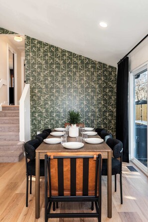 A dining area with a wooden table set for six, surrounded by black chairs. The room features a green patterned wallpaper and a large window with a view of the outdoors. - Arvada Retreat | Garage | Near Olde Town+Red Rocks (Arvada)