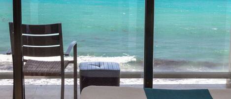 A room with a large window overlooking the ocean, a bed, a chair, and a small table.