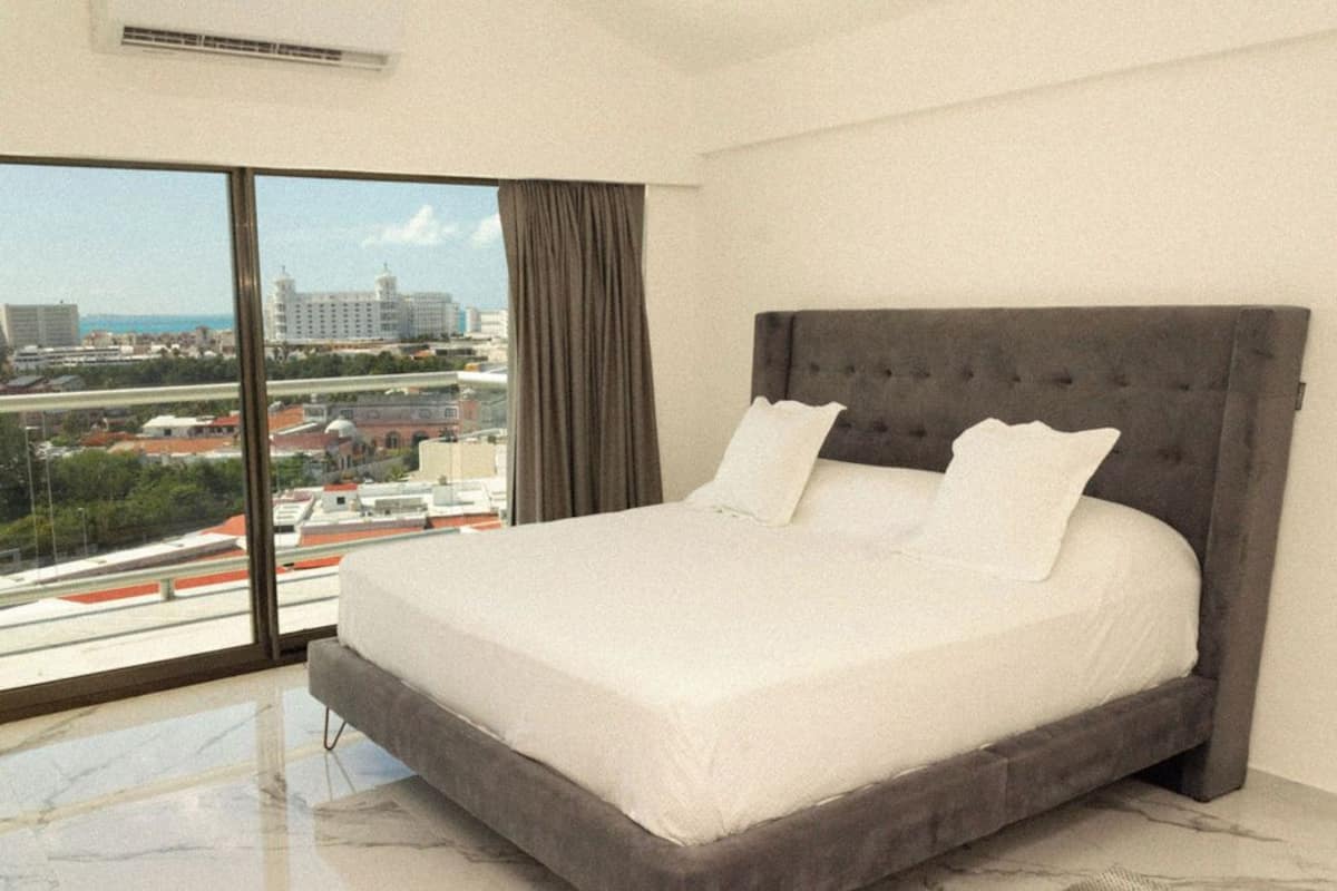 A modern bedroom with a large bed, a view of a cityscape through floor-to-ceiling windows, and a marble floor.