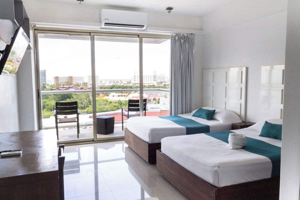 A hotel room with two beds, a desk, and a large sliding glass door leading to a balcony with a city view.