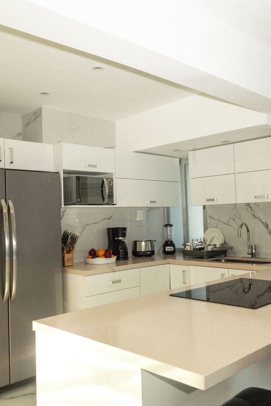 Una cucina moderna con mobili bianchi, un frigorifero in acciaio inossidabile, un forno a microonde, una macchina da caffè e un frullatore sul piano di lavoro.