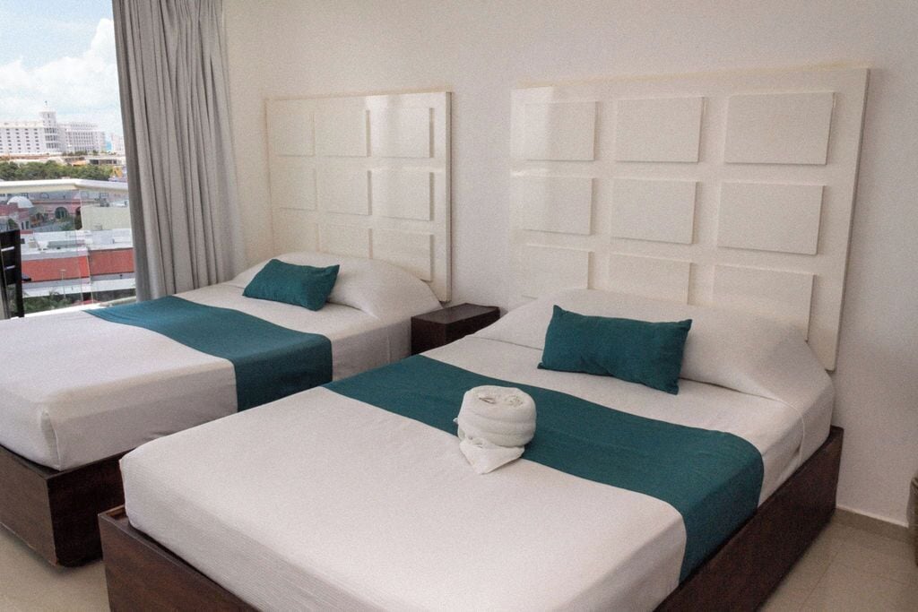 A hotel room with two beds, each with a white headboard and teal bedding. There is a window with curtains, and the room has a view of buildings outside.
