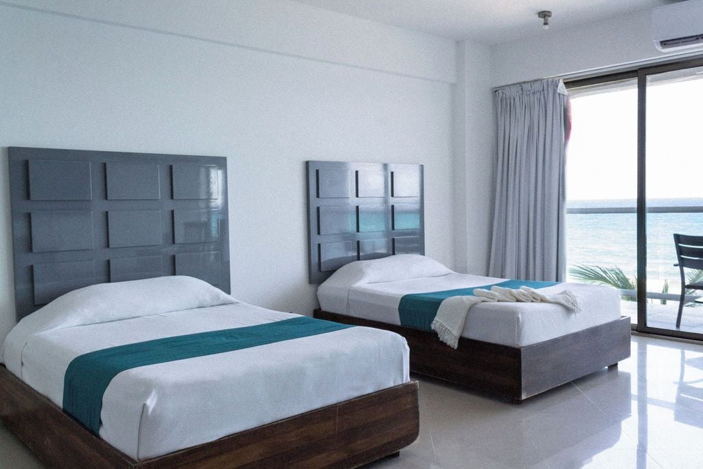 A hotel room with two beds, a large window with a view of the sea, and a balcony with a chair.