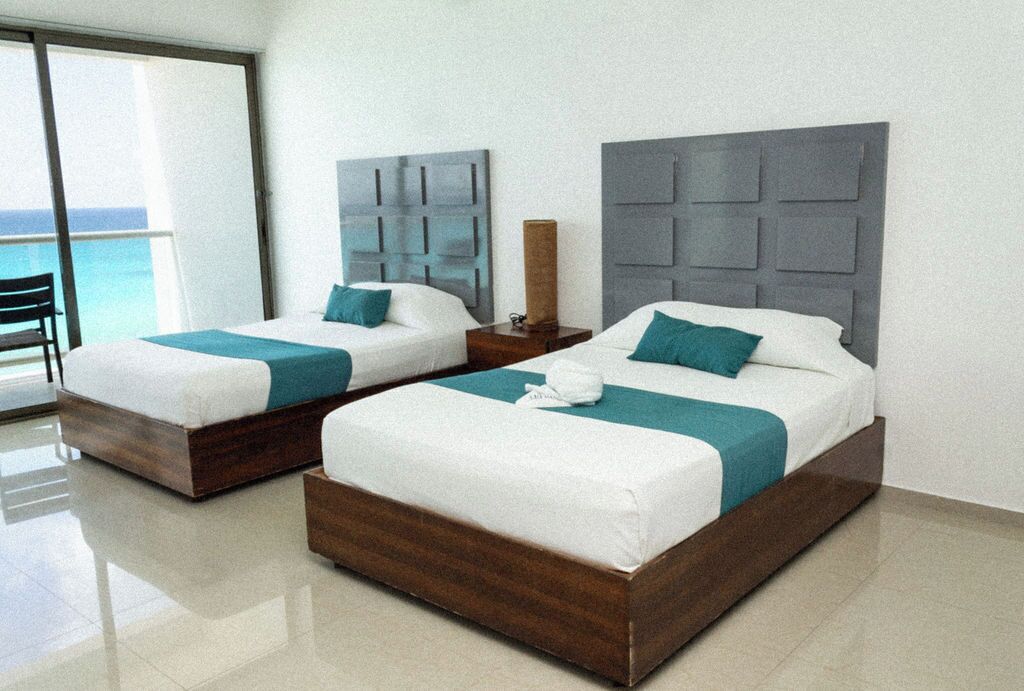 Two twin beds with wooden frames and teal bedding, a nightstand with a lamp, and a view of the ocean through a large window.