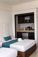 Habitación de hotel con dos camas, kitchenette y cabecera blanca.