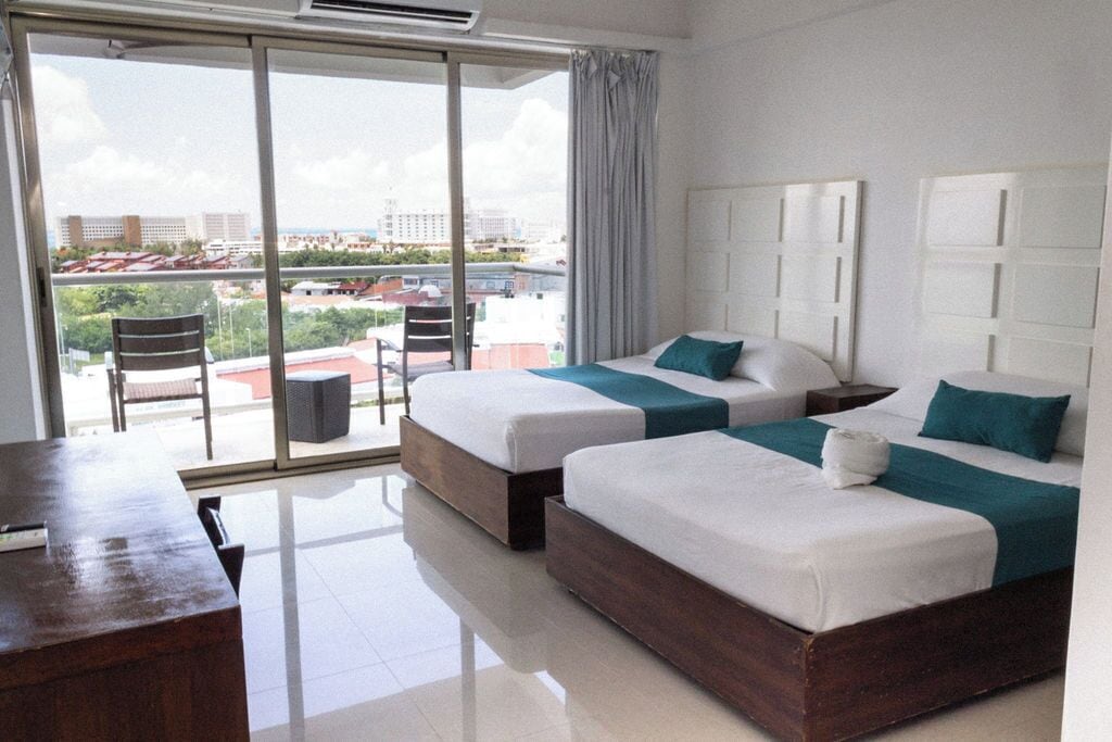 A hotel room with two beds, a desk, and a view of the city through large windows.