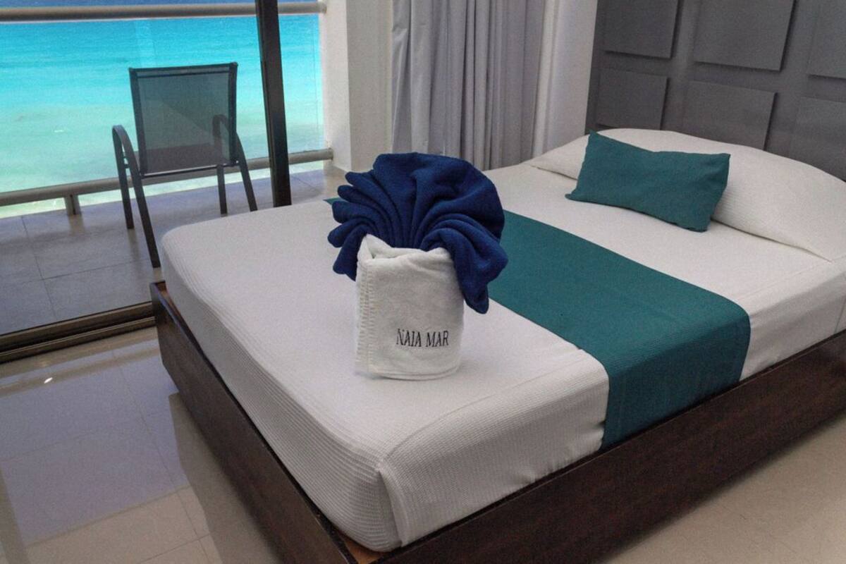 A hotel room with a bed, a view of the ocean, a chair, and a towel with "NAJA MAR" on it.