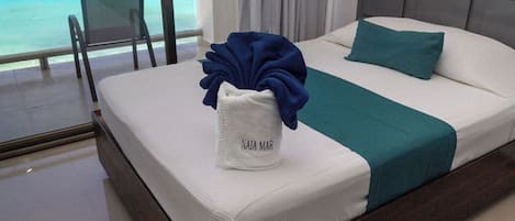 A hotel room with a bed, a view of the ocean, a chair, and a towel with "NAJA MAR" on it.