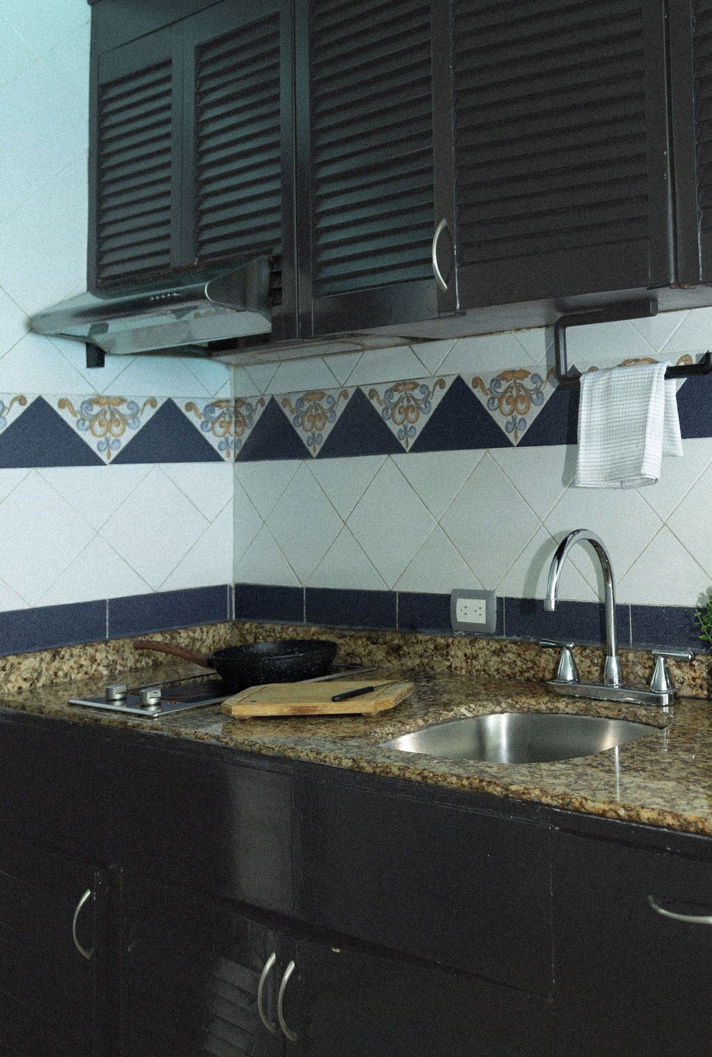 A kitchen with dark cabinets, a granite countertop, a stainless steel sink, and a backsplash with a geometric tile pattern.