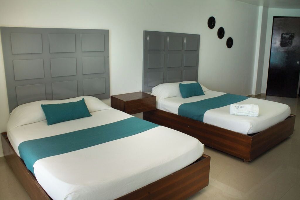 A hotel room with two beds, each with a wooden headboard and white bedding. There are two nightstands, one with a wooden finish and the other with a darker finish. The room has a neutral color scheme with a gray headboard and a white wall.