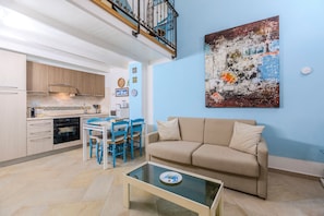 A living room with a beige sofa, a glass coffee table, and a blue dining area with chairs. - Castle Hill Loft Heart Cagliari by Yamar Villas (Cagliari)