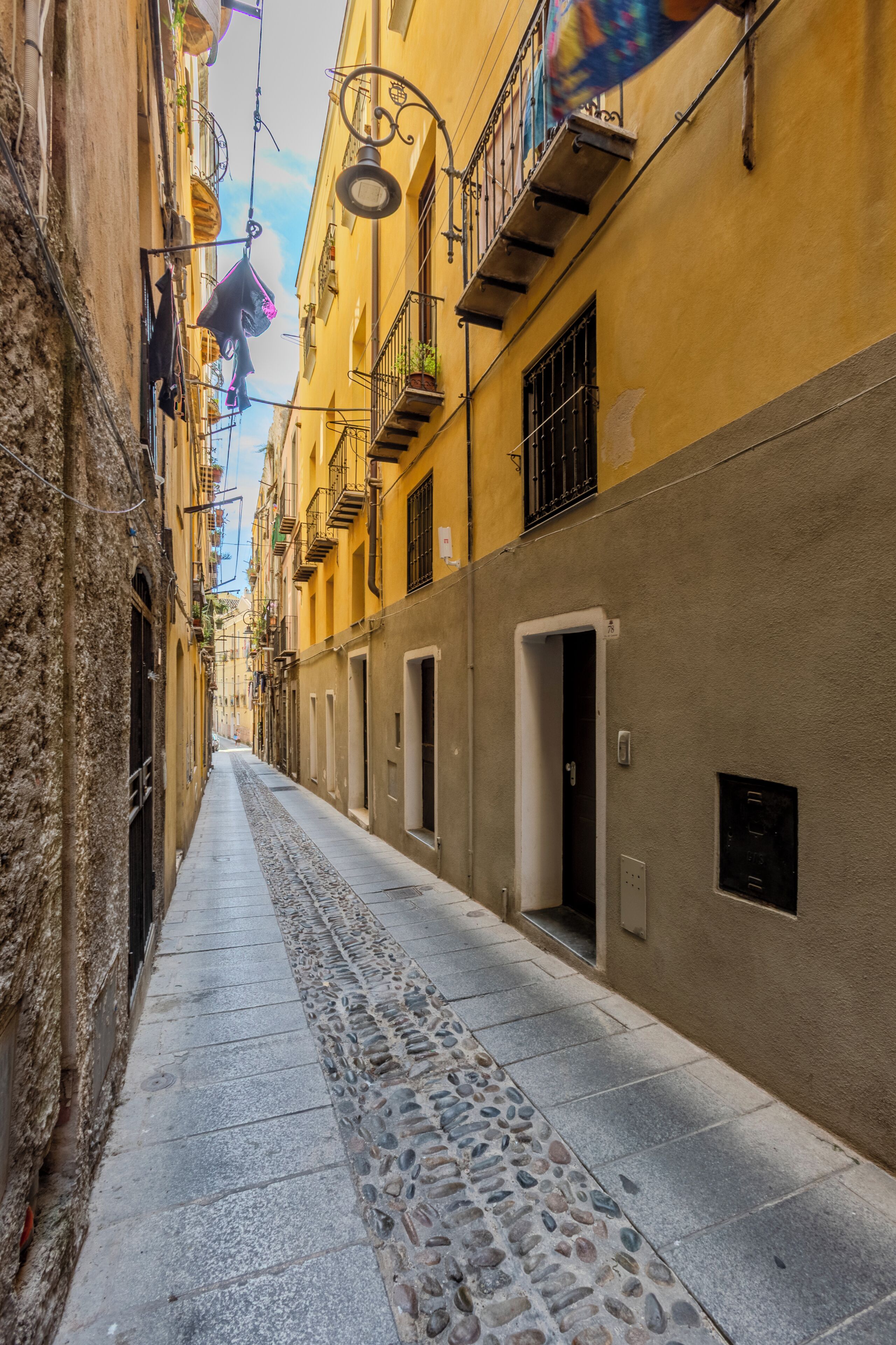 A narrow street with buildings on either side, featuring balconies with railings and potted plants.