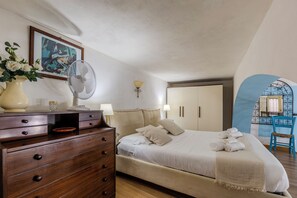 A bedroom with a bed, dresser, bedside lamps, a fan, a vase with flowers, a framed picture, and a blue chair. - Castle Hill Loft Heart Cagliari by Yamar Villas (Cagliari)