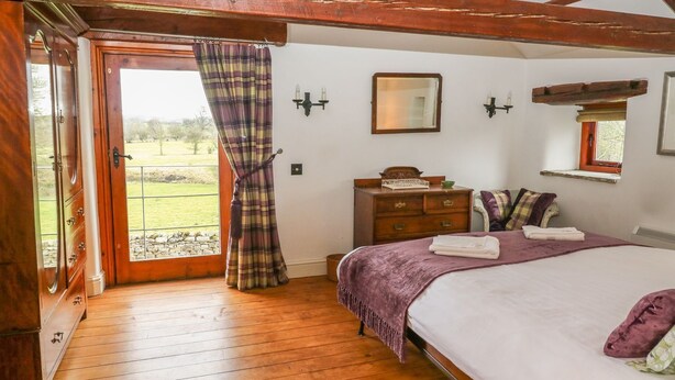 Een slaapkamer met een houten vloer, een bed met een paarse deken, een dressoir, een spiegel en een raam met uitzicht op de omgeving.