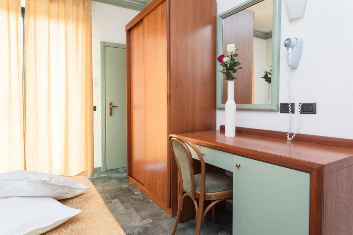 A bedroom with a wooden desk, a chair, a bed with pillows, a mirror, a vase with flowers, and a hairdryer on the wall.