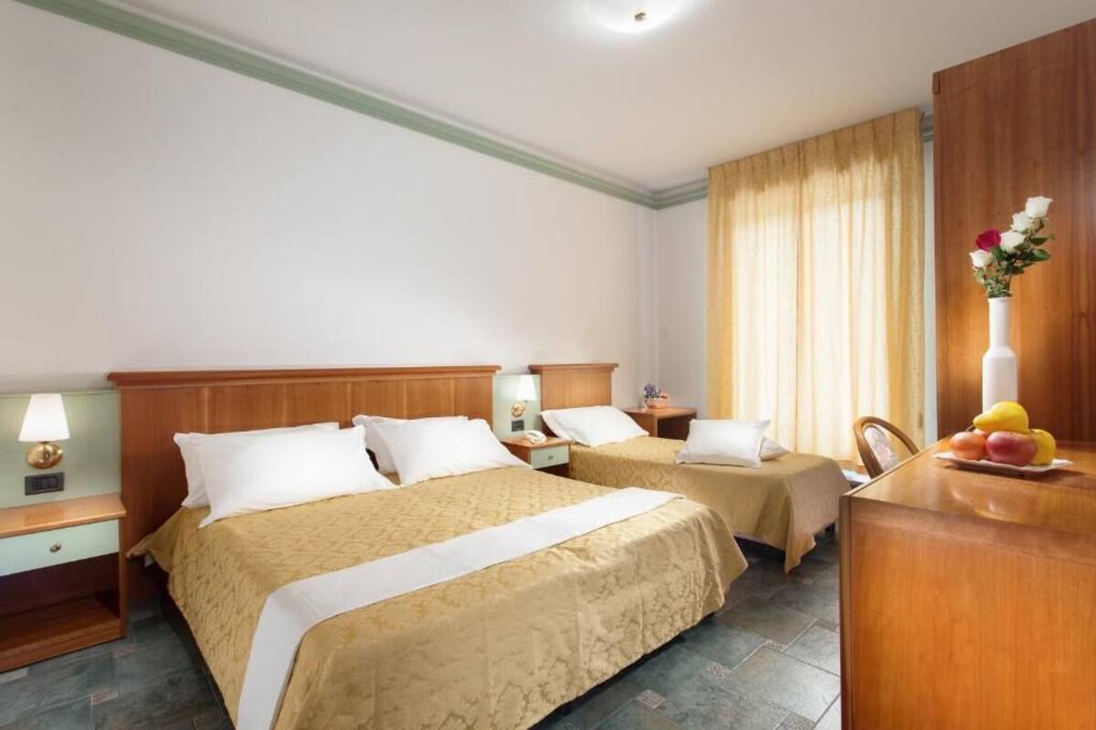 A hotel room with two beds, a wooden headboard, a desk with a vase of flowers, and a fruit bowl.