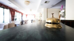Messinkinen kello tummalla työtasolla valoisassa huoneessa, jossa on suuret ikkunat ja punainen verho.