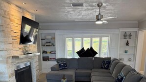 Un salon avec une cheminée, un canapé d’angle gris, une table basse et un ventilateur de plafond.